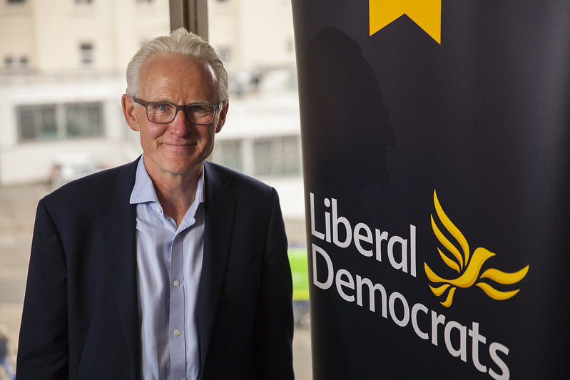 Sir Norman Lamb standing in front of the Liberal Democrats' party poster