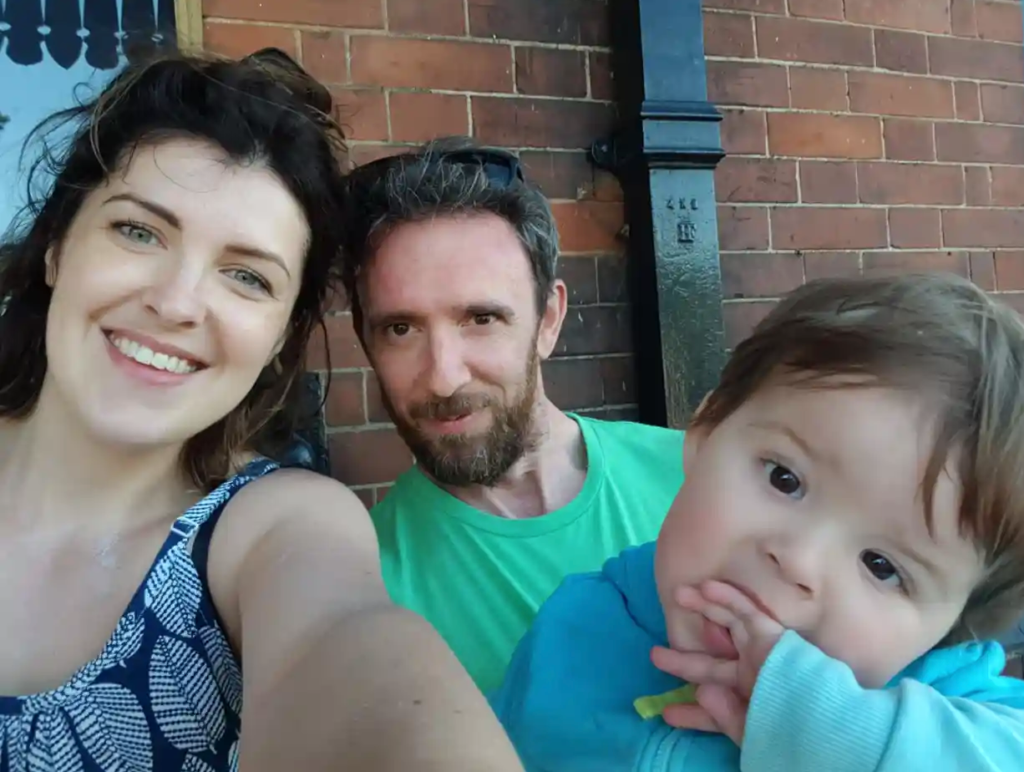 A selfie photography of two white adults, one male and one female smiling and holding a small male child
