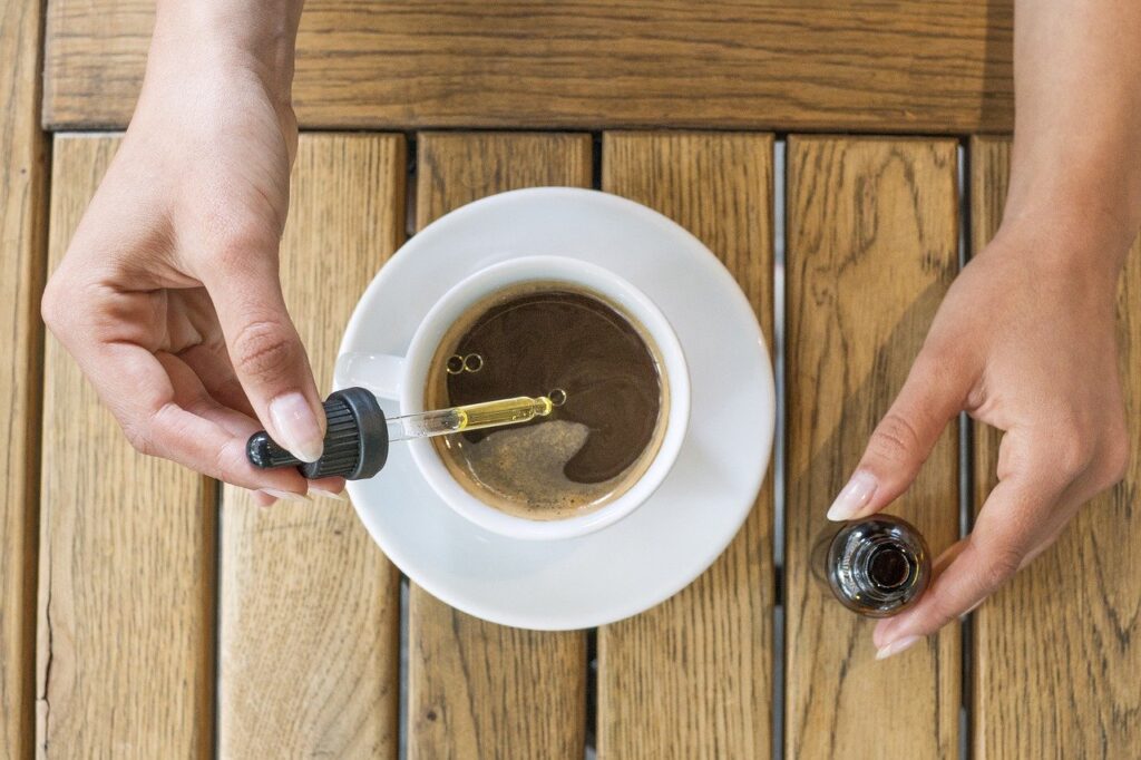 Two hands putting drops of a yellow CBD into a cup of coffee on a wooden table