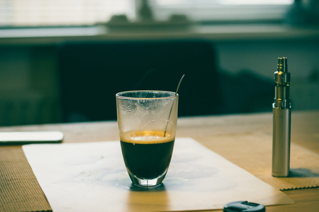 A vape device next to a small shot of coffee in a glass container with a spoon in it.