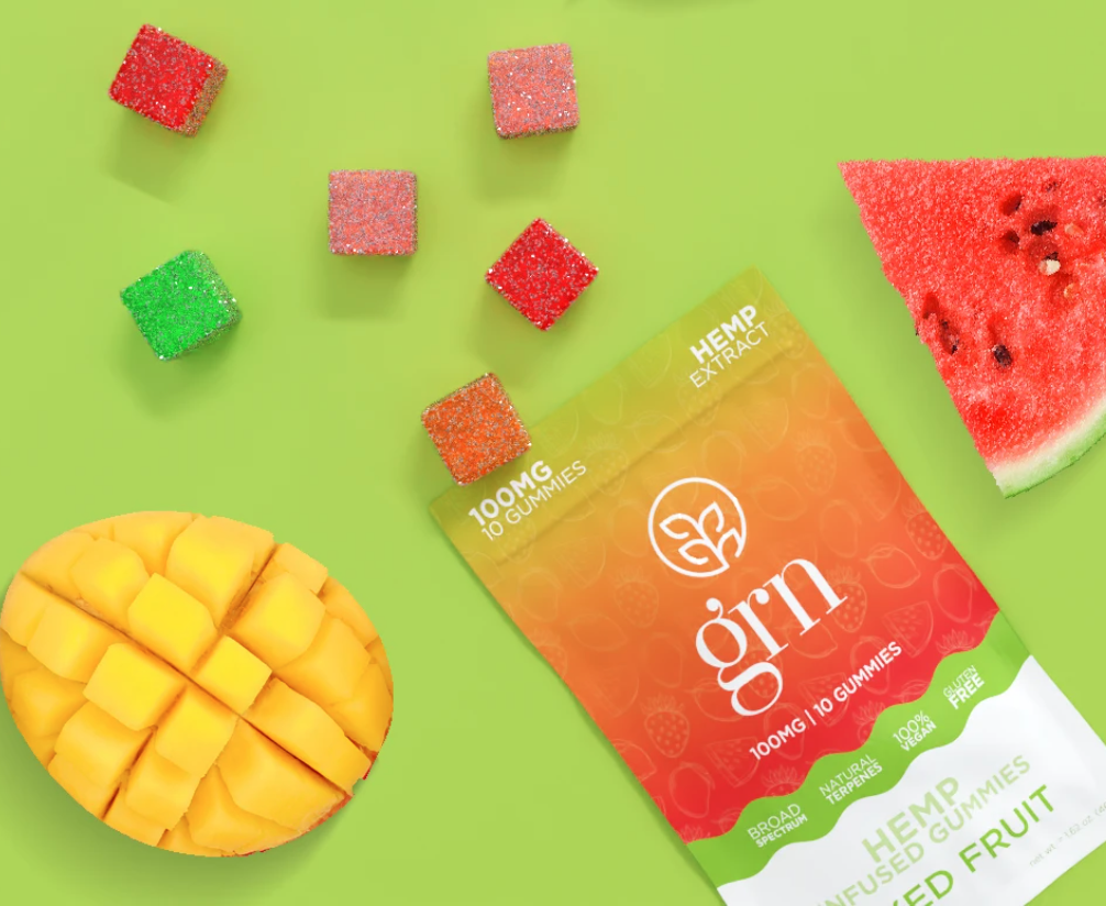 A packet of GRN CBD sweets on its side, open to reveal cubes of gummies. Slices of fruits are also beside the packet on a lime green background