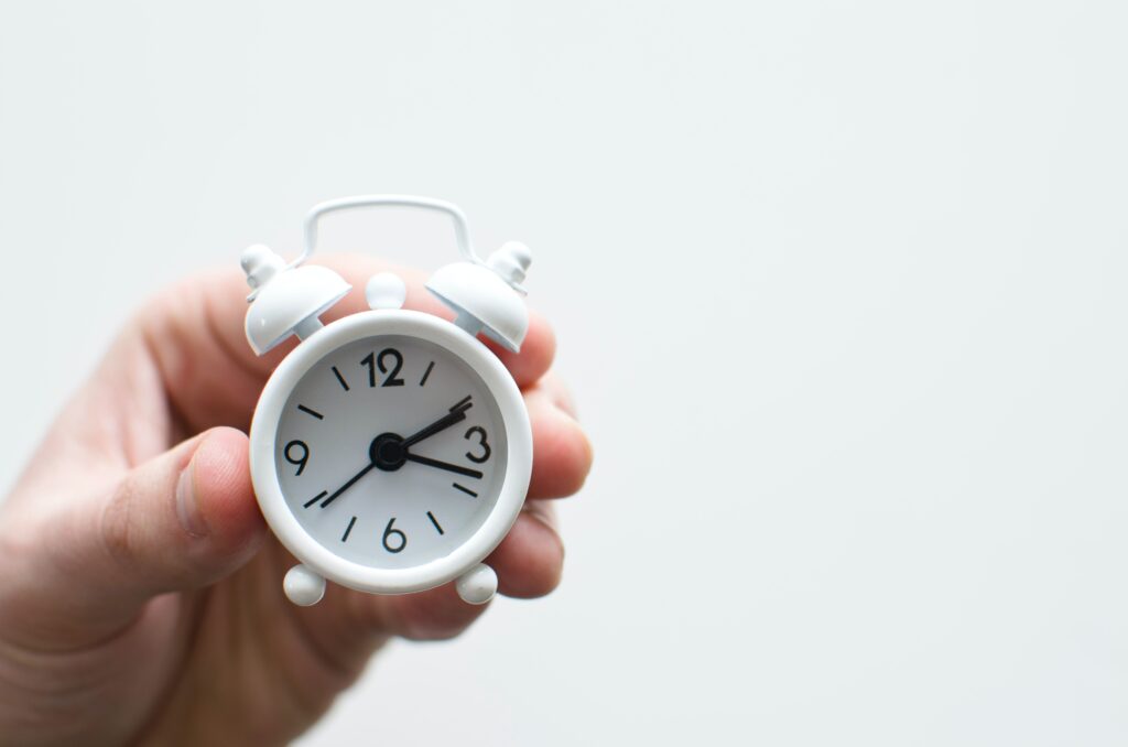 A hand holding a mini white clock which is showing the time as 2:20pm