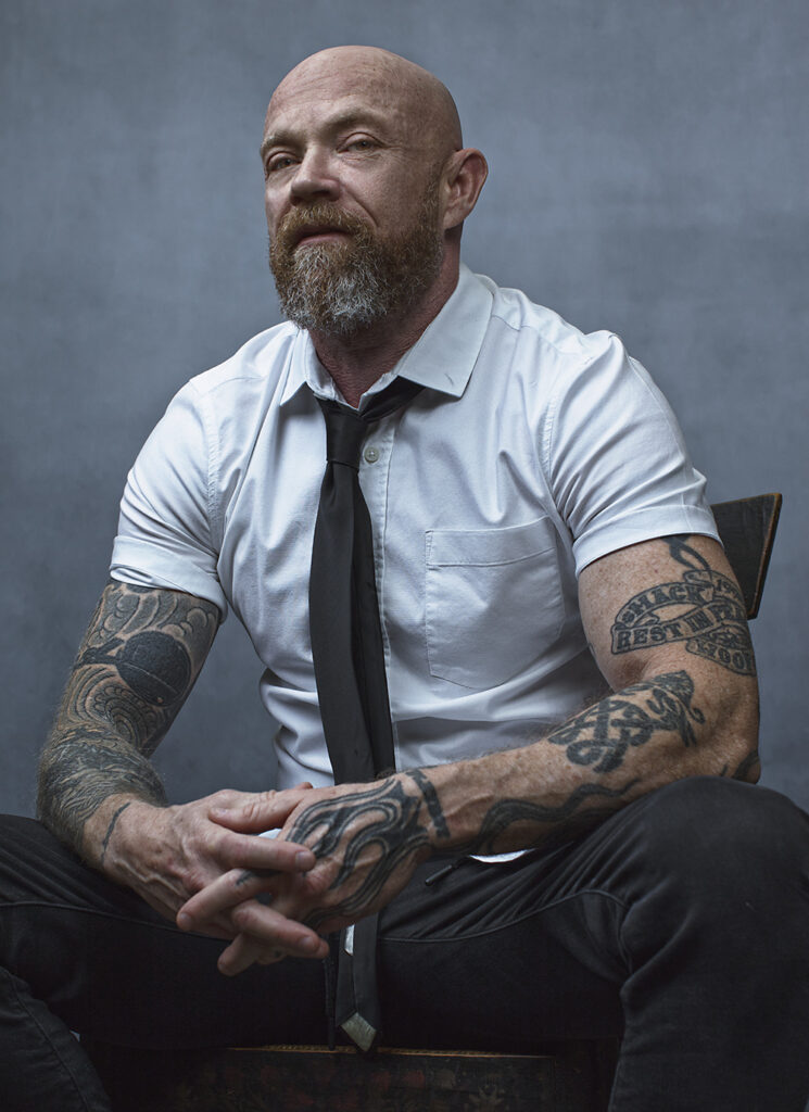 A man dressed in black and white,  bearing tattoos on his arms, is photographed while seated. 