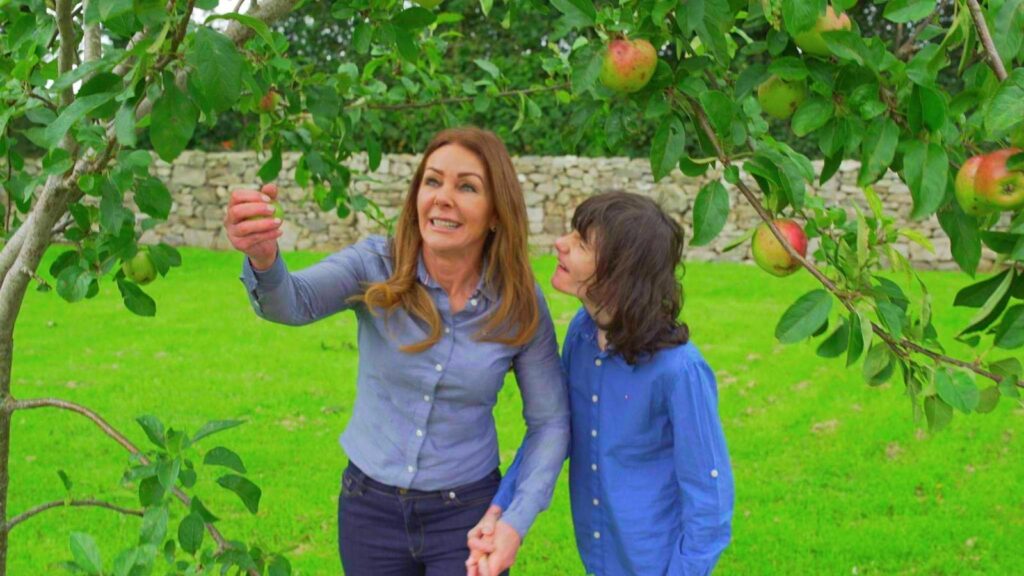 A mother and her son are pictured picking apples from an apple tree, wearing shades of blue. 