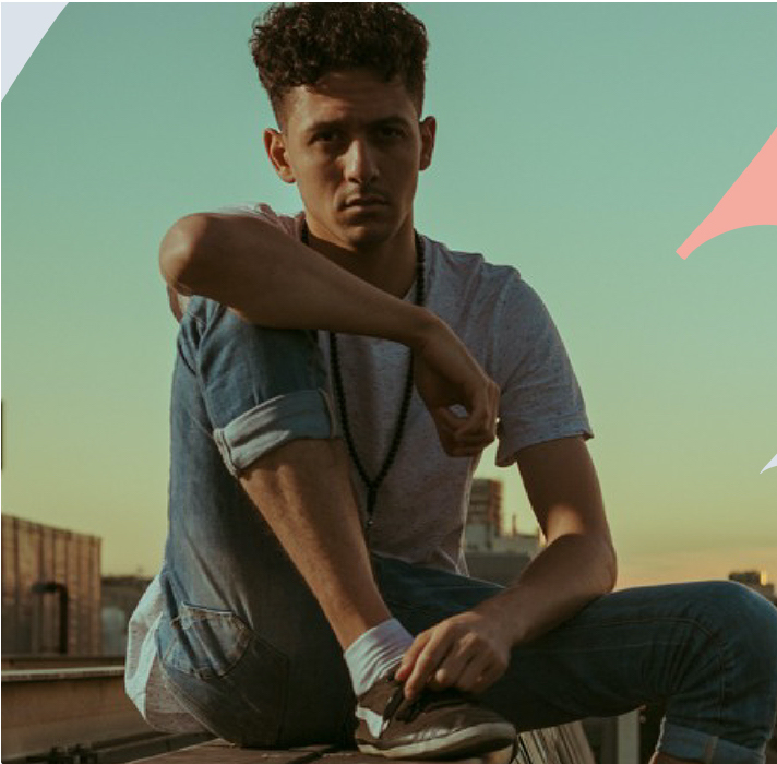 A man wearing denim and grey sits in a relaxed pose on a wall surrounded by blue sky. 