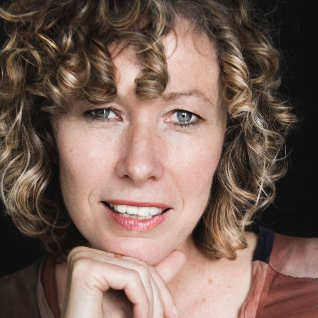 A woman with short, curly blonde hair and blue eyes is pictured smiling at the camera. 