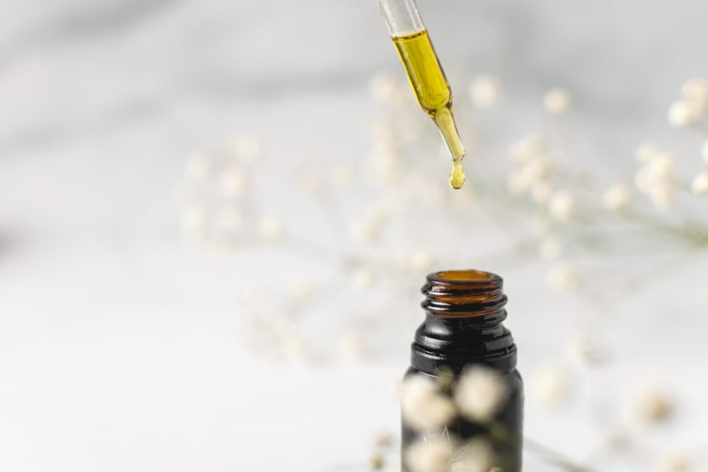 A dropper with oil poised above a brown glass bottle. There are white circular flowers dotted around the bottle.