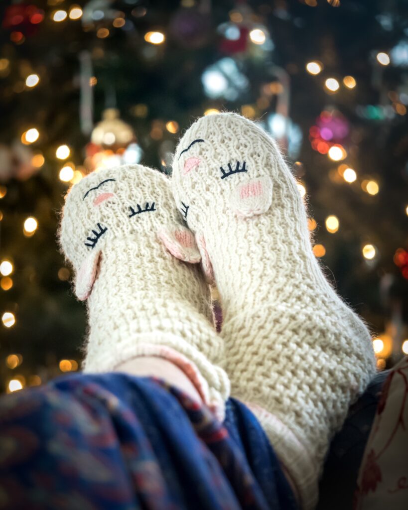 Two feet resting on a sofa with knitted sheep socks on. There is a twinkly xmas tree the background shining.