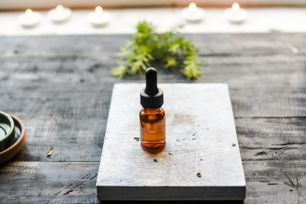 A small brown bottle of oil on a wooden chopping. 
there are also herbs in the background.