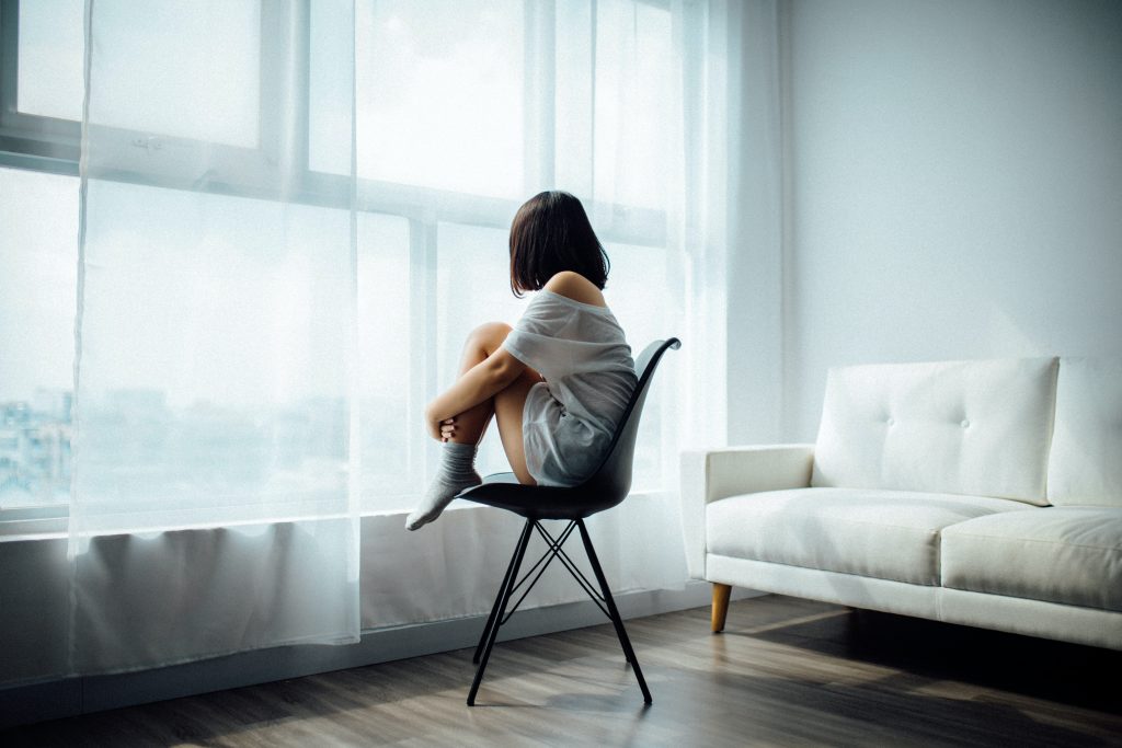 A woman with dark hair sits on a chair near a large window covered with voils. A white sofa is placed the right of the image