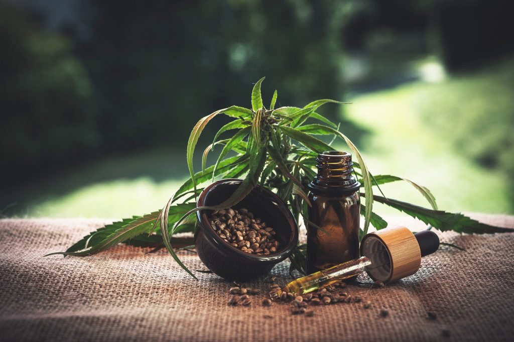 A brown bottle of oil sits on a hemp blanket with a cannabis leaf on top. There is a small brown bowl of hemp seeds to the left and a dropper containing on oil on the right