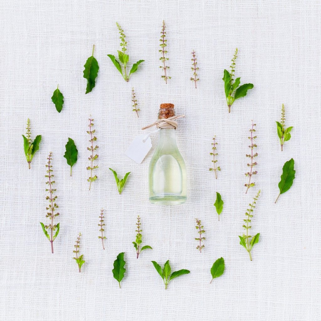 A collection of herbs, leaves and flowers on a white background surrounding a small bottle of oil with a wooden stopper. There is a string bow around the neck of the bottle.