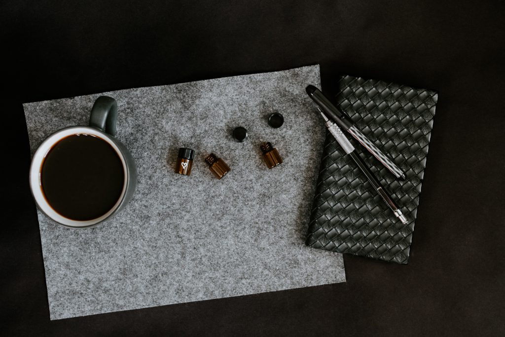 A black background that has. a vape pen, a diary, a coffee cup and small oil bottle on it