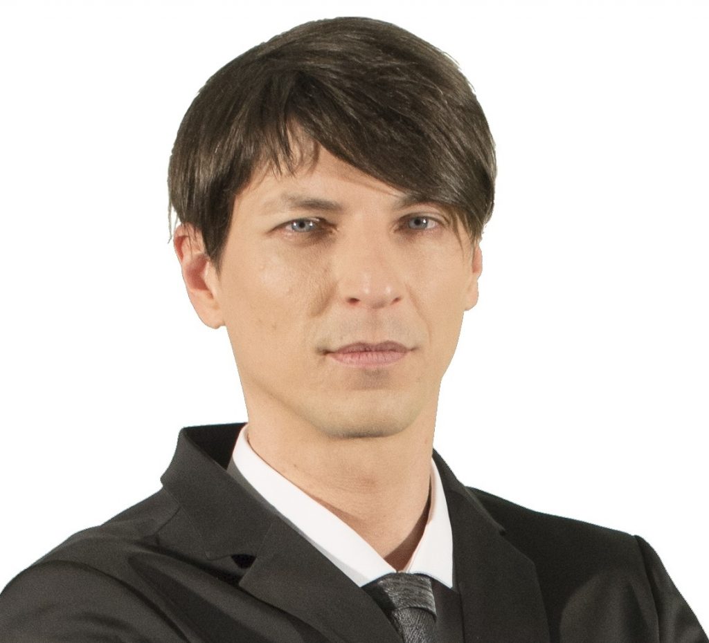 A man with black hair wearing a black suit and tie posts for a photograph 