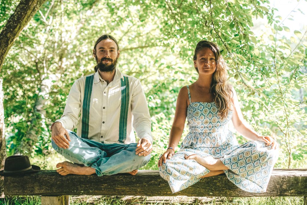 A man and woman sit cross-legged in front of greenery 