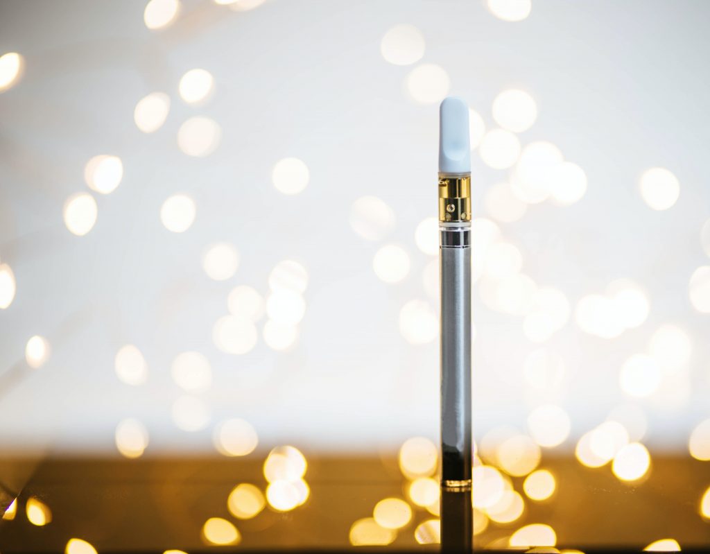 A thin silver and white cannabis vape with a background of white lights.