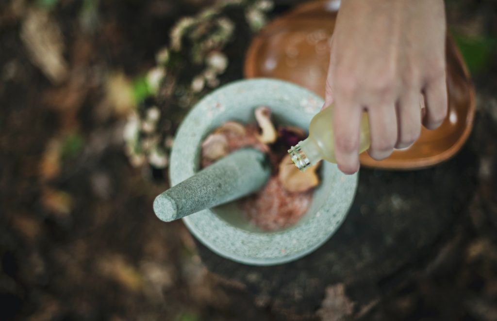 A hand pouring oil into a mortar and pestle full of flowers over a woodland ground.