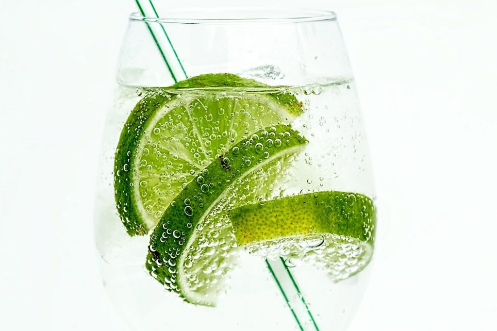 A clear glass of gin and tonic with a straw and three slices of limes in it