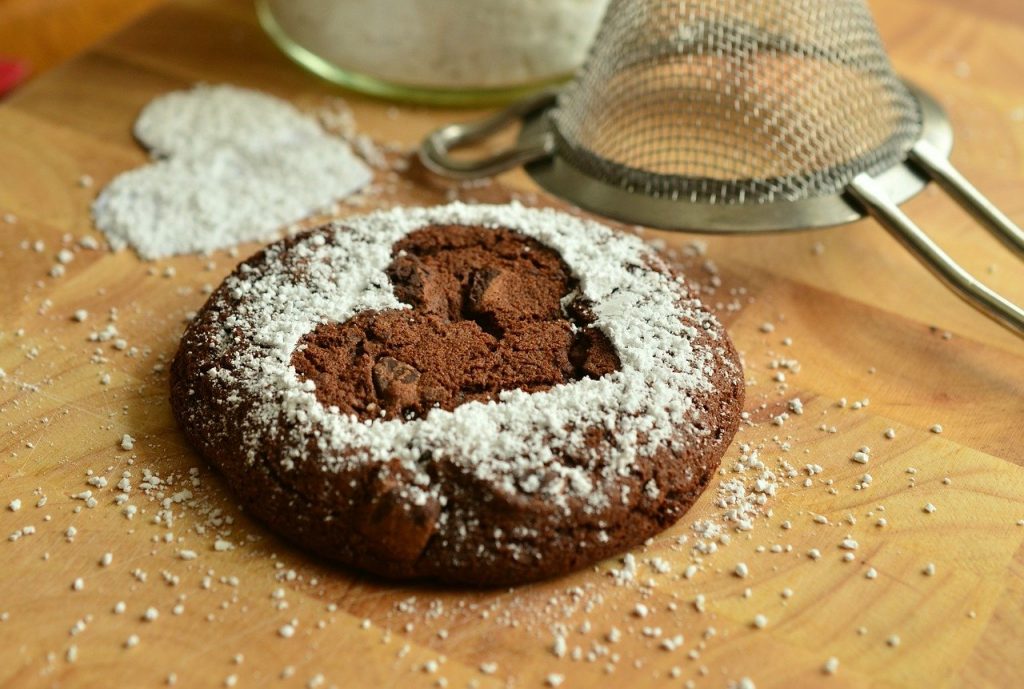 A chocolate brownie on the desk with a heart cut out of white sieved flour on top. The sieve is also lying to one side of the edible.