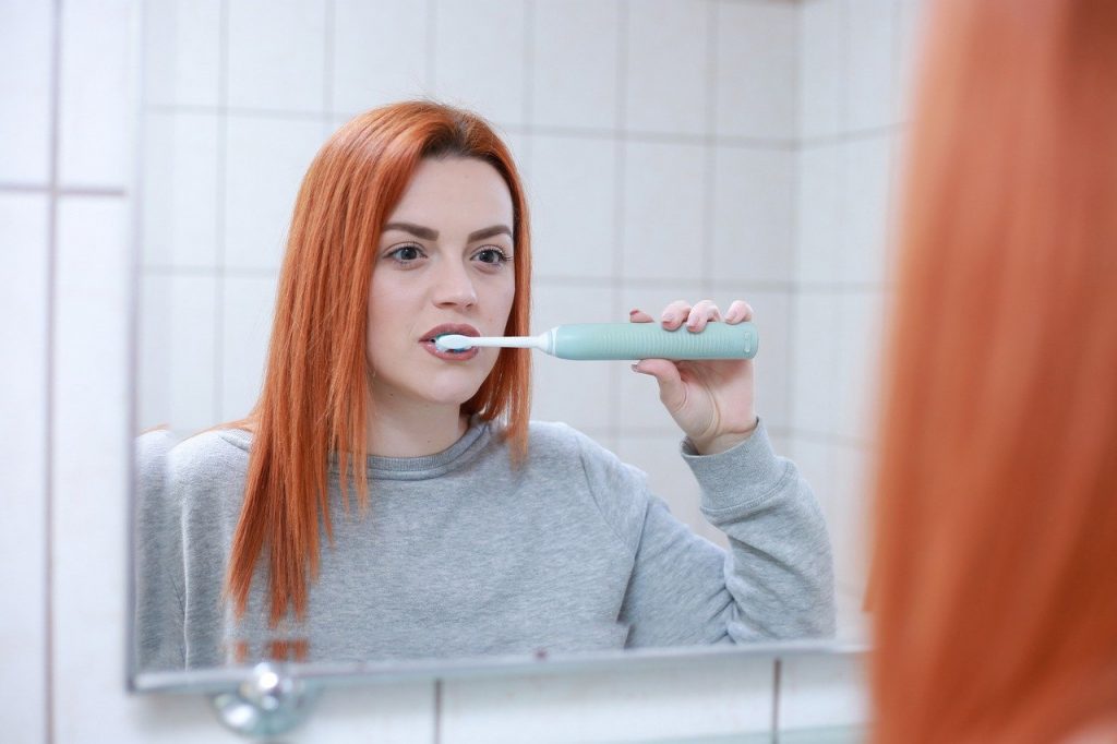 A white woman with bright red hair brushing her teeth with an aqua toothbrush in a white tiled bathroom