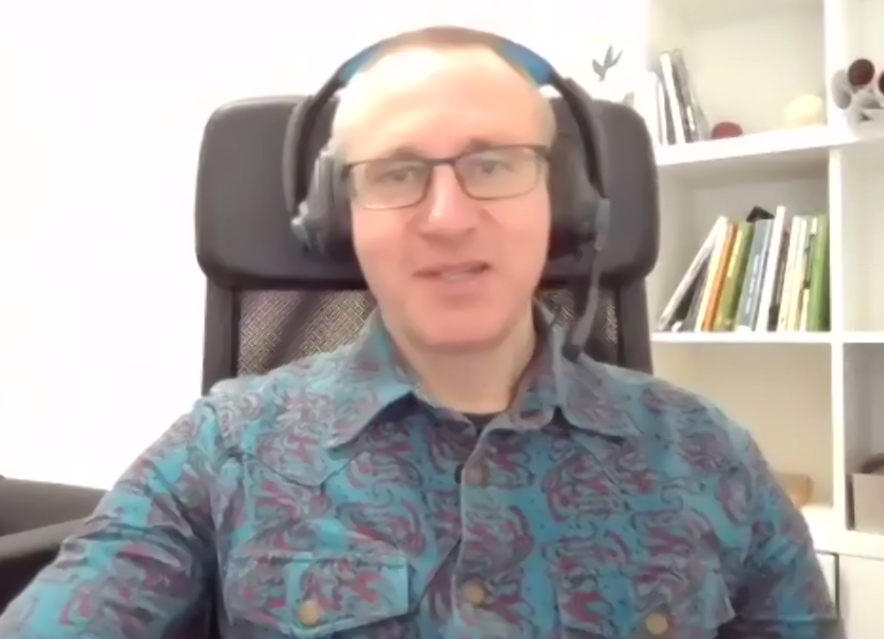 A man sits on a chair facing a screen on a zoom call. He is wearing a blue and purple shirt and headphones.