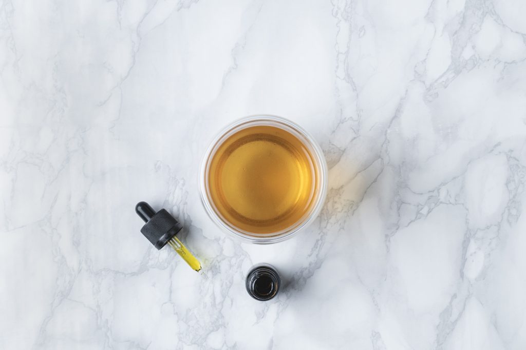 A marble counter top with a yellow glass cup of tea in it next to a brown CBD oil dropper and bottle